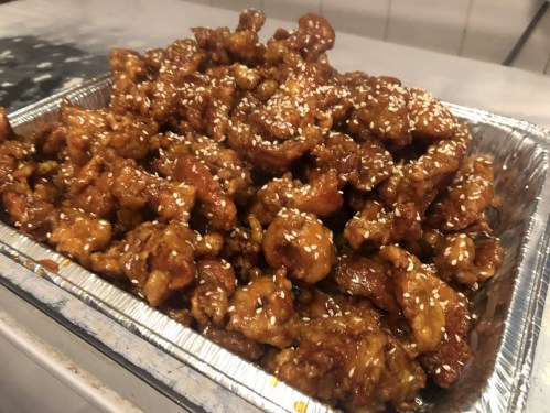 A large tray of crispy, golden-brown sesame chicken coated in a sweet sauce, garnished with sesame seeds.
