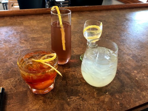 Four cocktails on a bar counter, each with a lemon twist garnish, showcasing different colors and glass styles.
