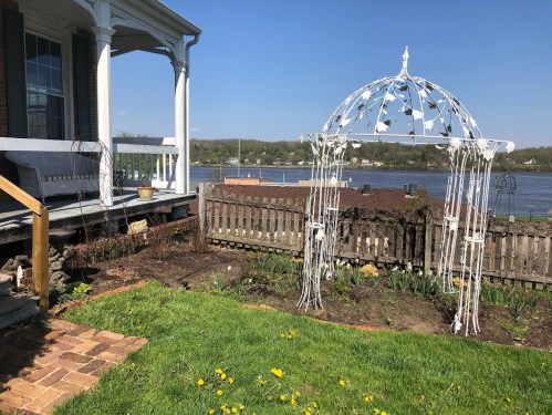 A white gazebo stands in a garden by a river, with a house and blue sky in the background. Green grass and flowers are visible.