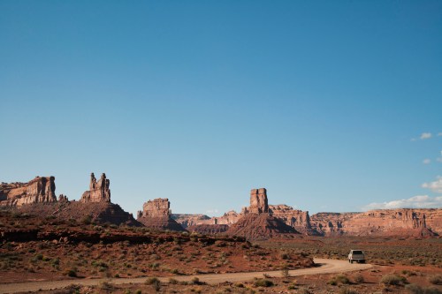 A scenic view of red rock formations under a clear blue sky, with a winding dirt road and a vehicle in the foreground.