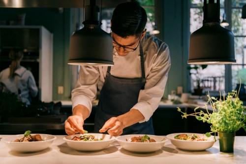 A chef carefully garnishes plates of food in a modern kitchen, with plants and dim lighting in the background.