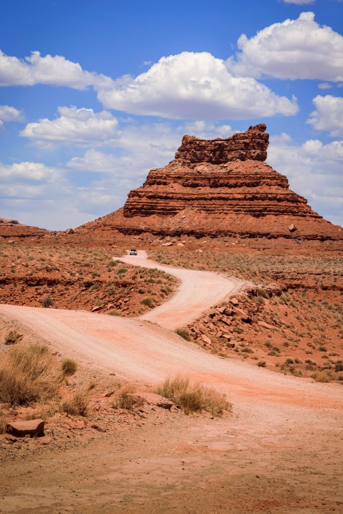 A winding dirt road leads to a rocky outcrop under a blue sky with fluffy clouds in a desert landscape.