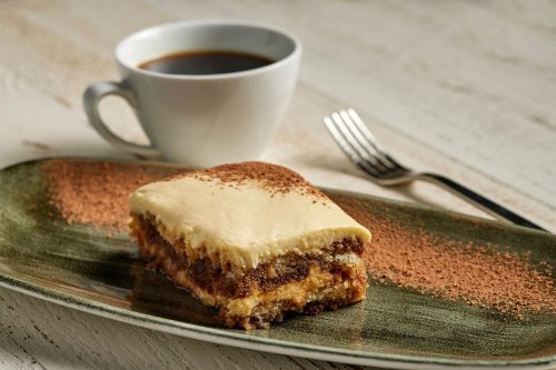 A slice of creamy dessert on a plate, dusted with cocoa powder, beside a cup of coffee and a fork.