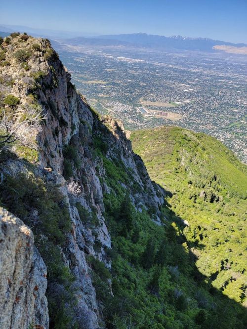 A rocky mountain ridge overlooks a lush green valley and distant mountains under a clear blue sky.