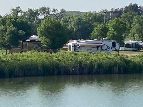 Two RVs parked near a lush green area by a calm lake, surrounded by trees and a clear blue sky.