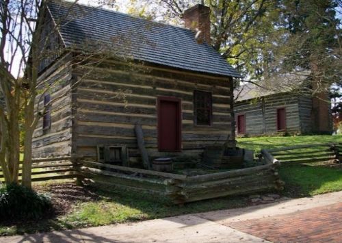 A rustic log cabin with a sloped roof, surrounded by greenery and a wooden fence, on a sunny day.