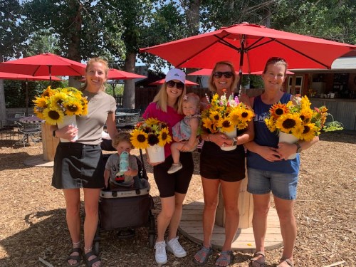 Five women stand together outdoors, each holding bouquets of sunflowers, with children and a stroller nearby.