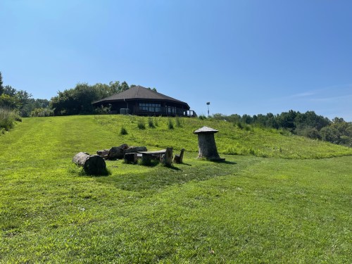 A grassy area with a wooden seating circle, a stump, and a building on a hill under a clear blue sky.