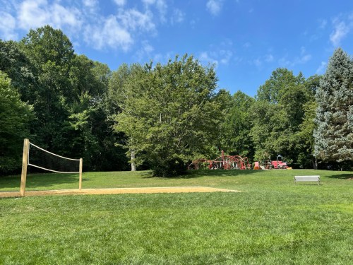 A sunny park scene featuring a volleyball net, grassy area, and playground equipment in the background surrounded by trees.