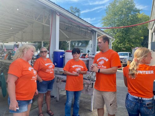 A group of five people in orange shirts chatting outdoors at a community event, with tents and vehicles in the background.
