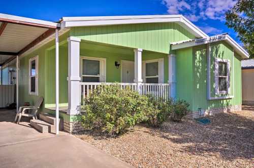 A green manufactured home with a front porch and shrubs, set against a blue sky with scattered clouds.