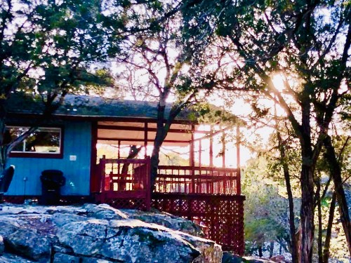 A cozy blue cabin with a red deck, surrounded by trees and rocks, illuminated by warm sunlight.
