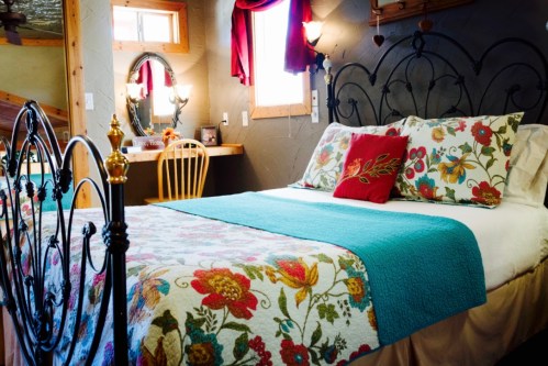 Cozy bedroom featuring a floral quilt, decorative pillows, and a wooden chair by a vanity mirror. Warm, inviting atmosphere.