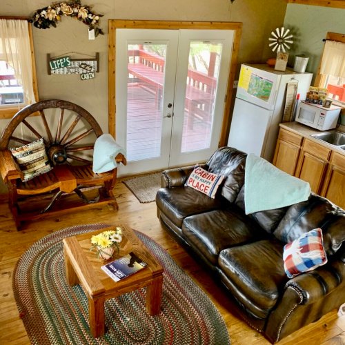 Cozy cabin interior with a leather couch, wooden wheel chair, coffee table, and a view of the deck through glass doors.