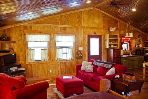 Cozy cabin interior with wooden walls, red furniture, and a kitchen area, featuring large windows and warm lighting.