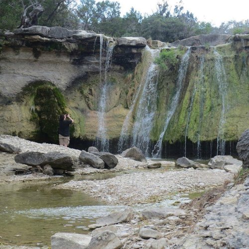 A person stands by a rocky stream, photographing a waterfall cascading over moss-covered rocks.