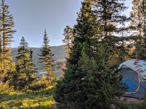A serene campsite by a lake, surrounded by tall trees and mountains under a clear blue sky.