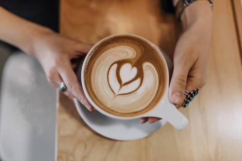 A person holding a cup of latte art with a heart design, set on a wooden table.