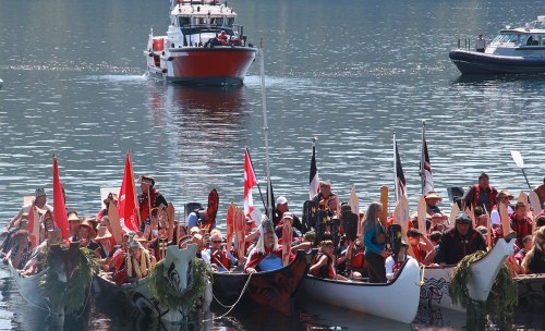 A group of people in traditional canoes paddling on a calm waterway, with boats and greenery in the background.
