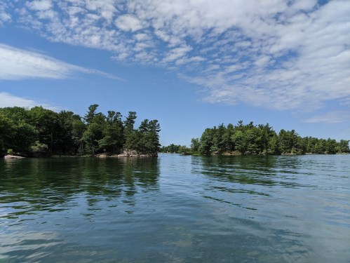 A serene lake scene with calm waters, surrounded by lush green trees and a blue sky with scattered clouds.
