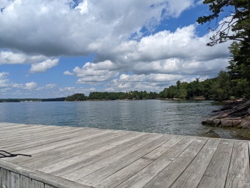 A wooden dock extends over calm water, surrounded by lush greenery and a partly cloudy sky.