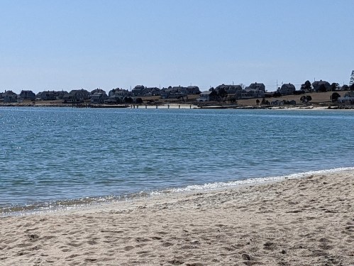 A calm beach scene with clear water, sandy shore, and distant houses along the coastline under a blue sky.