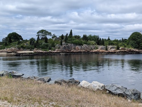 A calm body of water reflects a rocky shoreline and lush greenery under a cloudy sky.