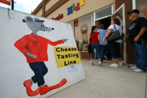 A cartoon mouse in a red shirt points to a sign reading "Cheese Tasting Line" outside a store with a line of people.