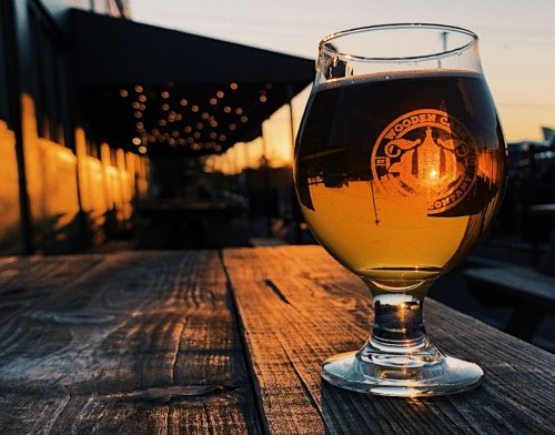 A glass of beer sits on a wooden table, with warm sunset light and string lights in the background.