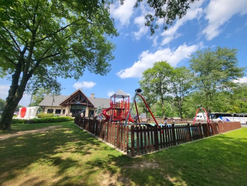 A playground with red equipment surrounded by a wooden fence, set in a green park under a blue sky with clouds.