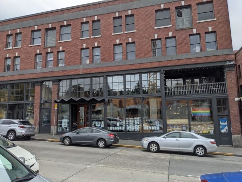A brick building with large windows, displaying colorful items inside, and parked cars along the street.