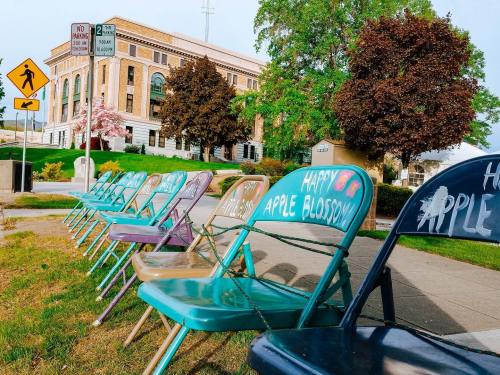 Colorful chairs lined up on a sidewalk, with "Happy Apple Blossom" painted on some, near a historic building and trees.