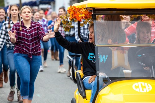 A woman waves from a yellow golf cart decorated with autumn leaves, while a group of people walks in a parade.