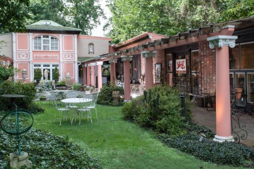 A charming garden with white tables and chairs, surrounded by lush greenery and elegant architecture.