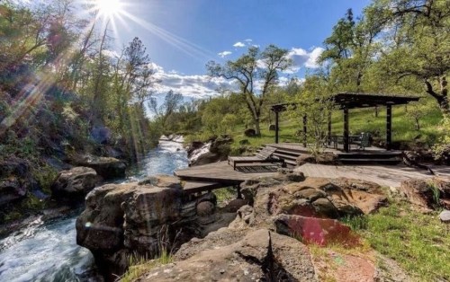 A serene riverside scene with sunlight, lush greenery, and a wooden deck by the water's edge.