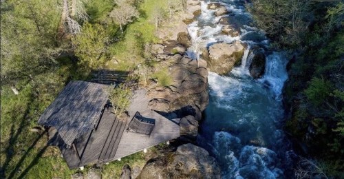 Aerial view of a wooden deck by a rushing river, surrounded by lush greenery and rocky terrain.