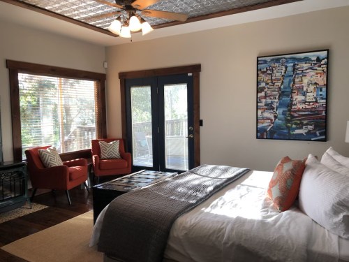 Cozy bedroom with a large bed, two red chairs, and a view of a deck through glass doors. Artwork on the wall.