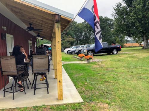 Outdoor seating area at a rustic building, with people chatting and a flag flying, surrounded by parked cars and flowers.