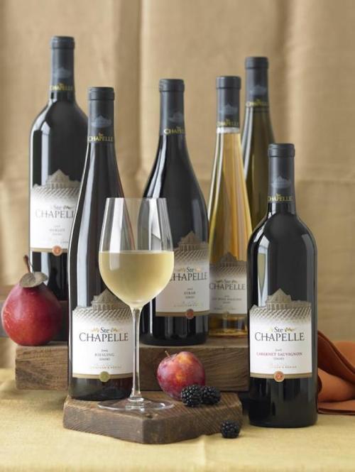 A selection of six wine bottles with a glass of white wine, surrounded by fruits on a wooden display.