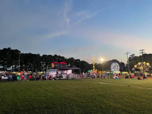 A lively outdoor fair scene at dusk, featuring a stage, rides, and a crowd enjoying the festivities on a grassy field.