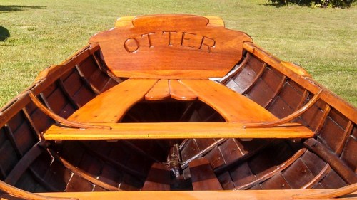 A wooden boat interior with the name "OTTER" carved on the backrest, featuring polished wood and a grassy background.