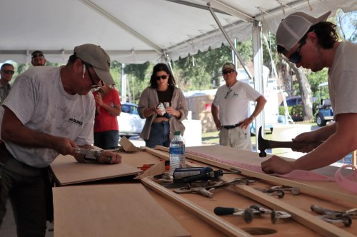 Two men work on woodworking projects under a tent, while a small crowd observes in the background. Tools are scattered on the table.