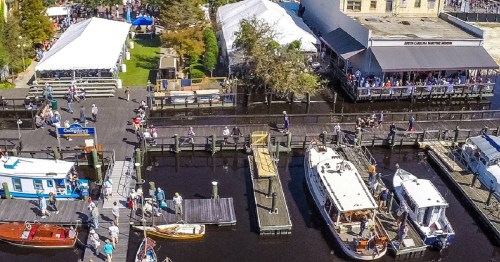 Aerial view of a busy waterfront with boats, people walking, and tents set up along the docks.