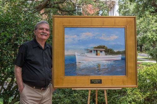 A man stands beside a framed painting of a white boat on water, surrounded by greenery.