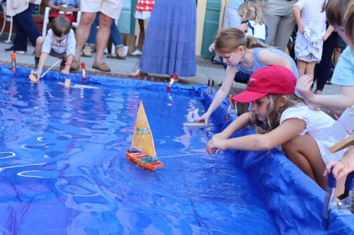 Children play with toy sailboats in a blue pool, focused on guiding their boats as others watch nearby.