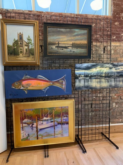 A display of five framed paintings on a wire grid, featuring landscapes, a fish, and a marina scene.