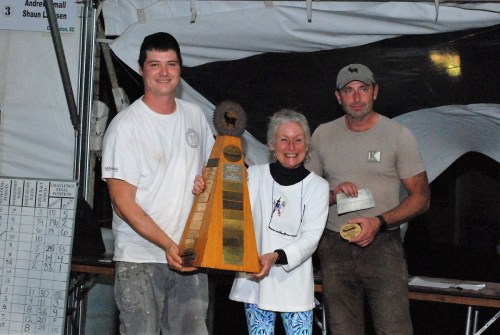 Three people stand together under a tent, holding a large trophy and awards, smiling at the camera.
