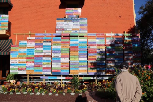 A person stands in front of a colorful wall of sponsor signs, surrounded by flowers and greenery.