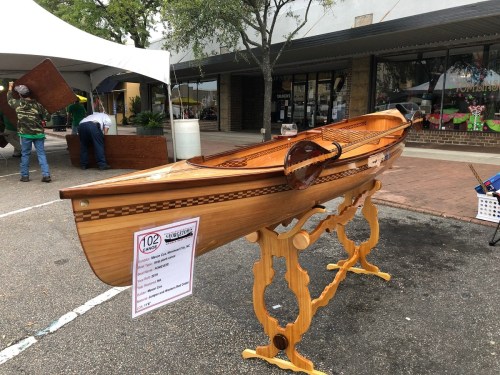 A beautifully crafted wooden kayak displayed on a stand at an outdoor event, with people and tents in the background.