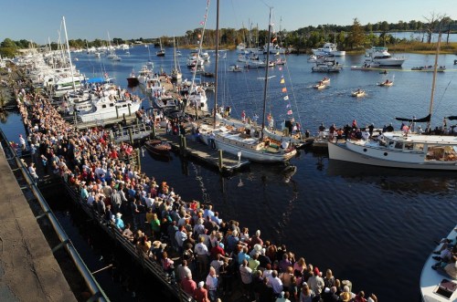 A large crowd gathers along a waterfront, with boats and yachts docked in the water under clear skies.